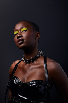 Woman with dark skin wearing bold yellow eyeshadow and a studded choker against a dark background.