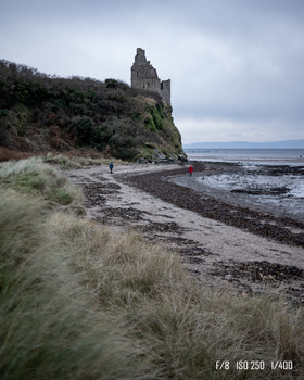 Medieval castle ruin on a grassy coastal cliff above a rocky beach with a wrecked boat.