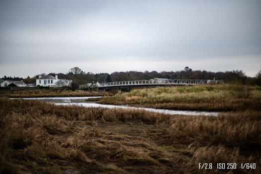 Coastal marshland with a winding creek, distant houses and a long boardwalk structure under overcast sky.