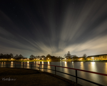 Nighttime waterfront scene with dramatic sky rays above illuminated buildings reflected in still water.