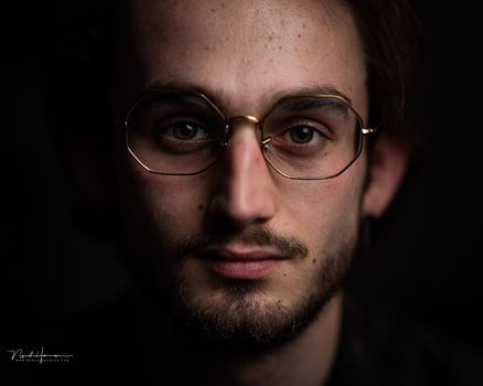 Man wearing round metal-framed glasses against dark background.