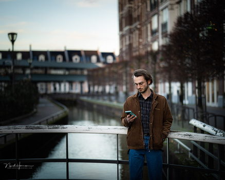 Man in brown jacket and blue jeans holding phone on urban bridge overlooking canal and historic buildings.