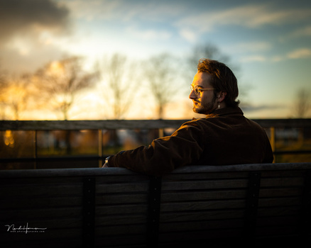 Man in sunglasses seated on a bench, gazing toward a golden-hour landscape with bare trees and a moody sky.