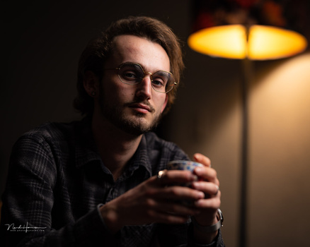 Man wearing glasses and dark plaid shirt gesturing while speaking, illuminated by warm overhead lighting.
