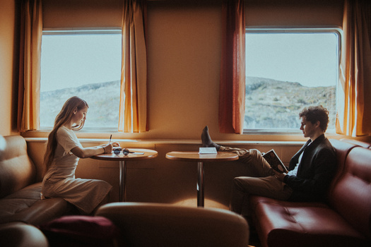 Two people seated across from each other at a small table beside large windows overlooking a mountainous desert landscape.