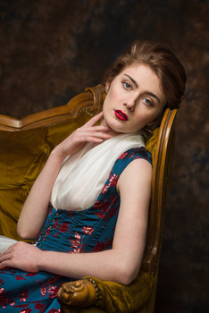 Woman in vintage-style dress seated in golden chair against dark textured backdrop.