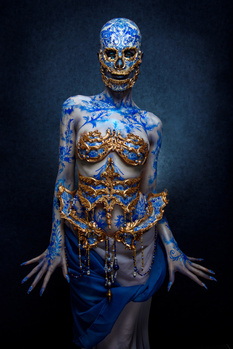 Body paint model with blue and gold ornamental designs covering torso, arms, and head against dark background.