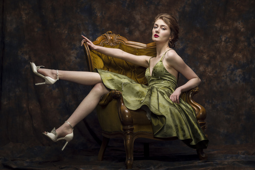 Woman in green dress seated on ornate chair, holding draped fabric with extended leg.