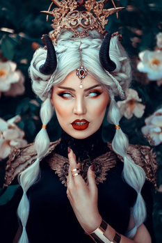 Person with pale blue hair, ornate crown, horns, and dramatic makeup against blurred floral background.