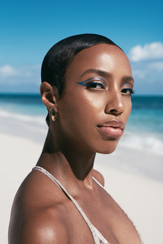 Woman with short dark hair and pearl earring looking toward camera at beach with ocean and sky behind her.