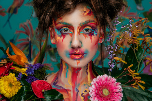 Person with vibrant multicolored face paint and dramatic eye makeup surrounded by fresh flowers against a teal background.