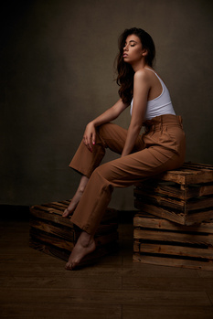 Woman in white tank top and tan pants seated on stacked books against neutral backdrop.