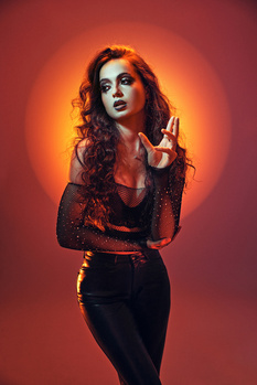 Woman with wavy red hair against warm orange backdrop, wearing dark fitted clothing and dramatic makeup.