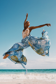 Woman jumping on beach with arms extended, wearing patterned blue fabric against clear sky and ocean.