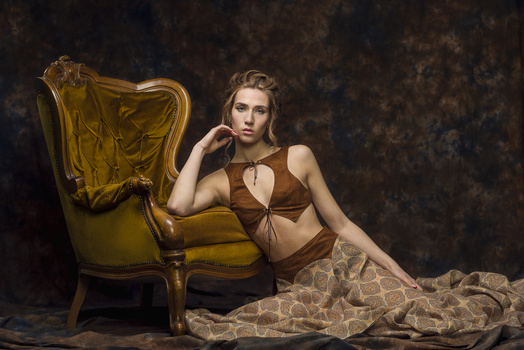 Woman in brown bikini top seated on ornate golden chair against textured dark backdrop.