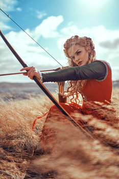 Woman drawing compound bow in desert landscape with warm golden light and blue sky.