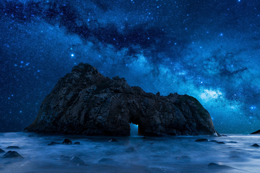 Rocky island silhouetted against a vibrant starry night sky with the Milky Way visible over calm ocean waters.