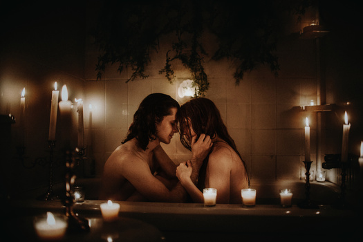 Two people in a candlelit bathtub, facing each other intimately surrounded by lit candles and greenery.