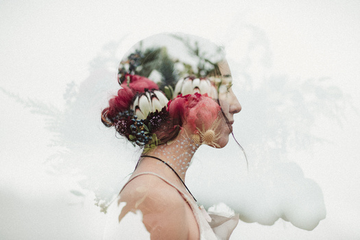 Side profile of woman's head filled with red flowers and foliage, blending into soft clouds.