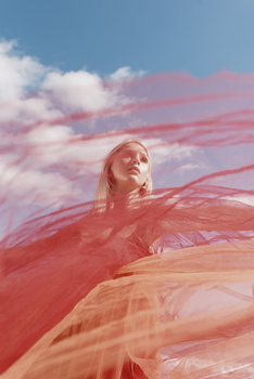 Woman looking upward with flowing red fabric against a blue sky with pink-tinted clouds.