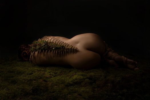 Artistic nude figure lying on moss-covered ground, dramatically lit against black background.