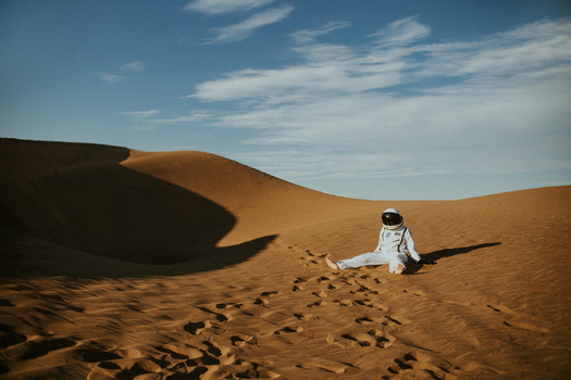 Astronaut in white spacesuit sitting on desert sand dune with footprints visible in foreground.