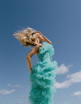 Woman in turquoise tulle dress with windswept blonde hair against clear blue sky.