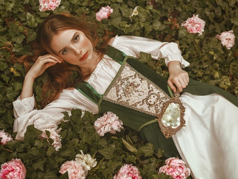 Woman in white blouse and gold-patterned skirt lying among pink roses and green foliage.