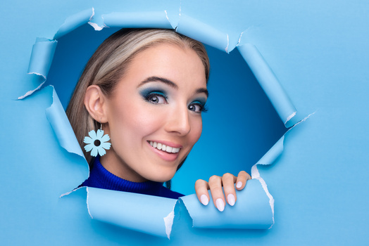 Young woman with blue eyeshadow peering through a torn blue paper hole, wearing white daisy earrings and smiling.