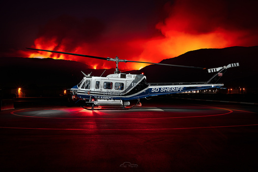 Sheriff helicopter illuminated by dramatic wildfire glow at night on landing pad.
