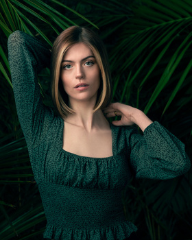 Woman in teal knit sweater posing against layered green palm fronds backdrop.