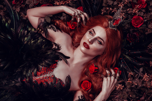 Woman with vibrant red hair reclining among black feathers and red roses in a dark, moody portrait.