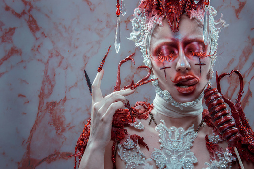 Portrait of figure with pale makeup, red eye designs, and elaborate white lace collar surrounded by red ornamental elements.