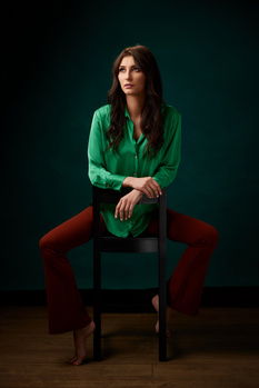 Woman in green shirt and burgundy pants sitting on wooden chair against teal backdrop.