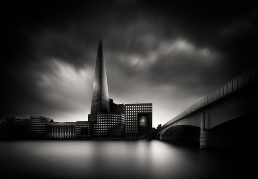 Modern cityscape with tall pointed tower and bridge reflected in still water under dramatic storm clouds.