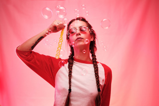 Person wearing round glasses and red cardigan blowing bubbles against pink background.
