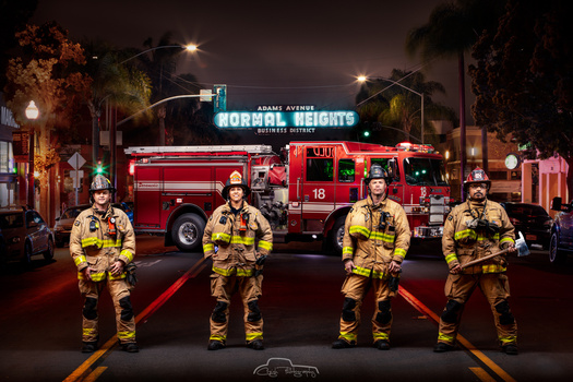 Four firefighters in yellow turnout gear standing in formation on a street at night with fire trucks and neon signage behind them.