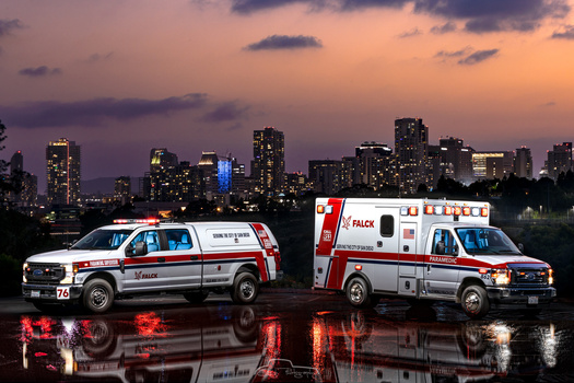 Two ambulances parked side by side at dusk with a city skyline and purple sky in the background.