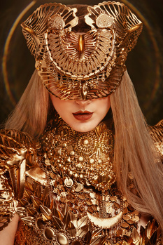 Model wearing ornate golden mask and sequined garment against dark background.