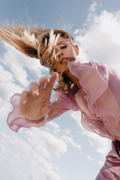 Woman in pink dress with windswept blonde hair against blue sky with white clouds.