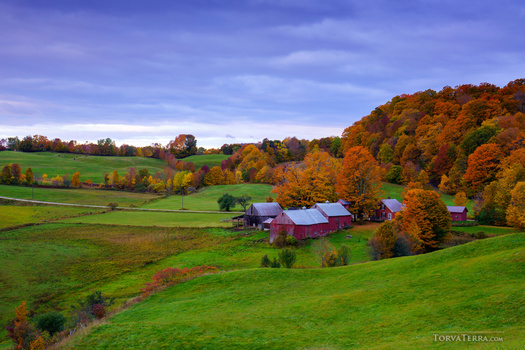 Red barn and farm buildings nestled among rolling green hills with golden autumn foliage.