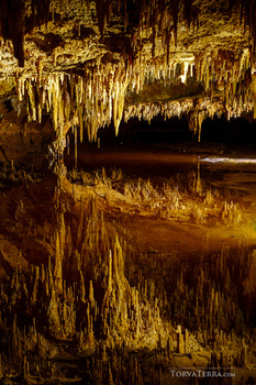 Underground cave chamber with stalactites and stalagmites illuminated in golden-yellow light, reflecting in still water below.
