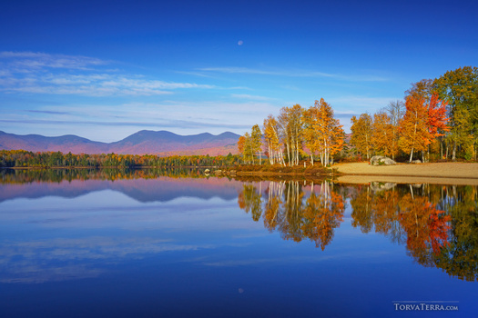 Autumn forest reflected in still lake water with distant mountains under clear blue sky.