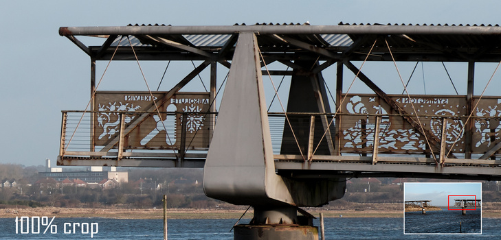 Industrial bridge structure with ornate metalwork panels suspended over water, shot from below against clear sky.