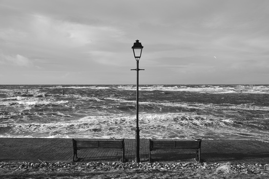 Empty bench and street lamp on a pebble beach facing a turbulent sea under overcast sky.