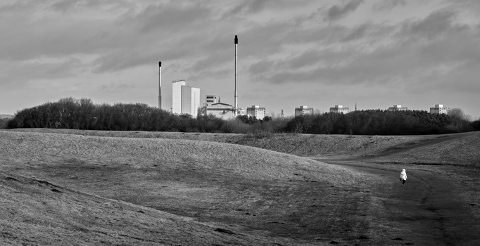 Industrial complex with tall smokestacks rises above bare rolling fields under overcast sky.