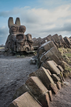 Large stone sculpture of a stylized animal head with geometric features overlooking a rocky breakwater.