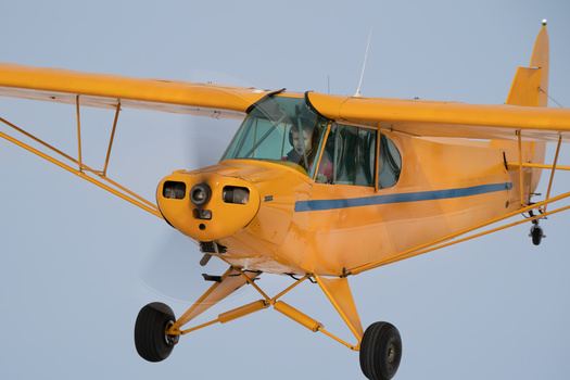 Yellow high-wing aircraft in flight against a clear blue sky, shot from below.