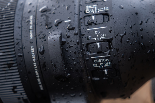 Close-up of a telephoto lens barrel covered in water droplets, showing focus ring and AF/MF and custom switches.