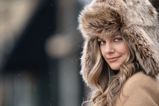 Woman wearing a fur-lined winter hat with long blonde hair, photographed outdoors in falling snow.